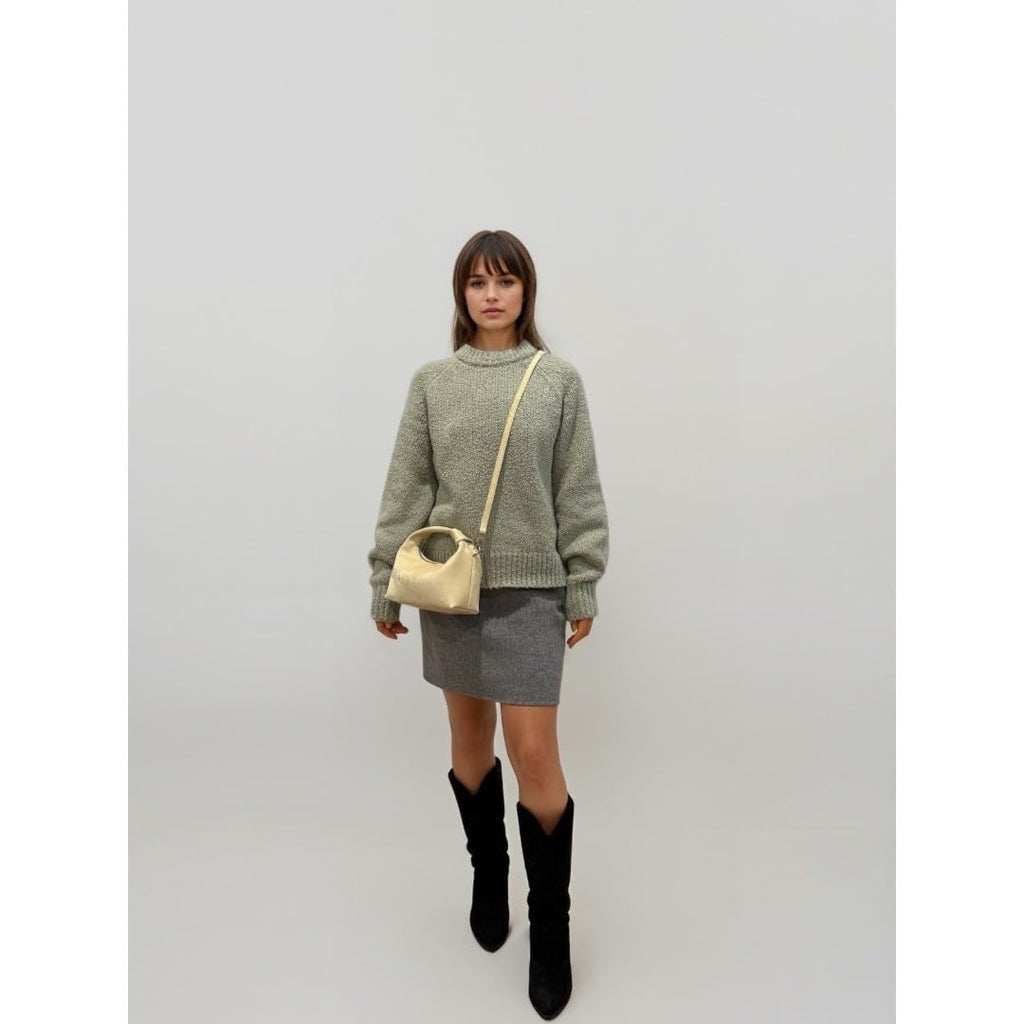 Woman wearing a green sweater, gray skirt, and black boots with a golden yellow handbag against a white background